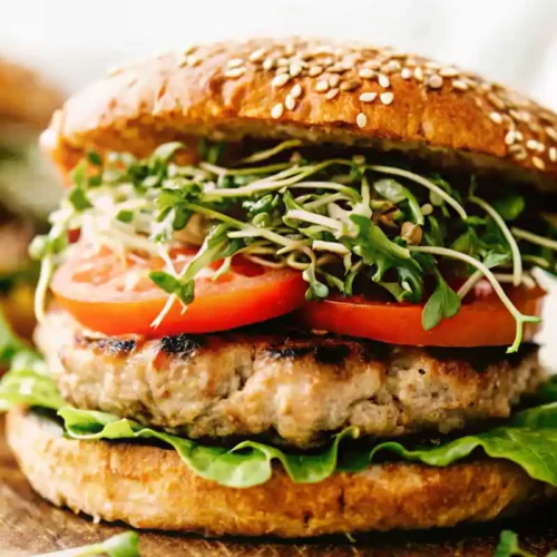 A stacked turkey burger on a sesame bun with fresh greens, sliced tomatoes, and sprouts on a rustic wooden board.