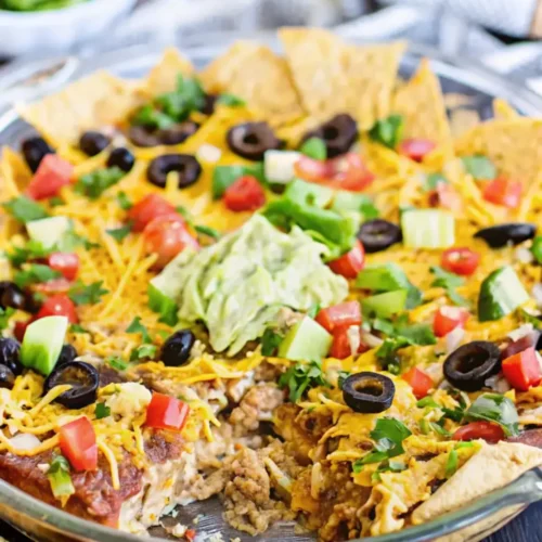 A layered taco dip topped with guacamole, black olives, and chopped tomatoes, served with tortilla chips.