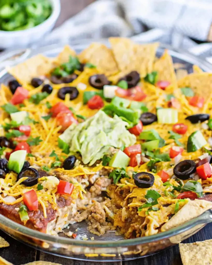 A layered taco dip topped with guacamole, black olives, and chopped tomatoes, served with tortilla chips.